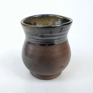 May include: A ceramic cup with a rounded body and a wide, flared rim. The lower portion features a matte, textured brown finish, while the upper section and interior are glazed in a glossy, dark charcoal black with subtle metallic highlights.