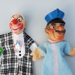 May include: Two hand puppets, one is a clown with a plaid jacket and a green saxophone, the other is a police officer with a blue uniform and a pink trim.