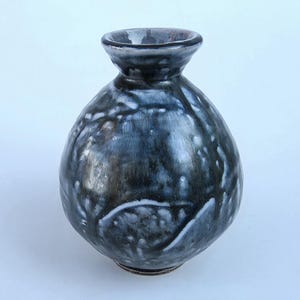 May include: A small, bulbous ceramic vase with a narrow neck and a flared rim. The surface features a glossy, mottled glaze in shades of deep charcoal, black, and white, creating a marbled effect with visible drips and textured highlights.