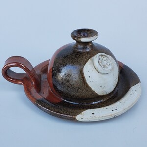 May include: A ceramic butter dish with a brown, white, and red glaze. The lid has a round knob with a decorative design.