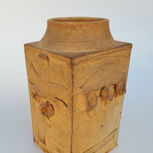 May include: A square ceramic vase with a textured, light brown finish. The vase has a wide mouth and a slightly flared rim. The surface of the vase is decorated with raised, irregular shapes.