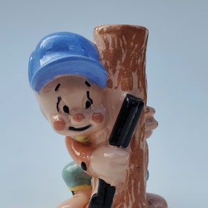 May include: A ceramic figurine of a cartoon boy wearing a blue baseball cap, holding a rifle and hiding behind a tree trunk. The figurine is painted in shades of brown, blue, green, and yellow.