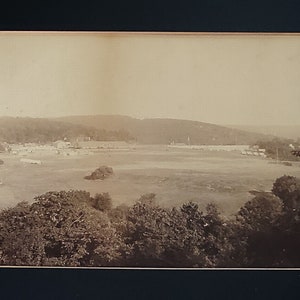 May include: A black and white photograph of a large, open field with a small town in the background. The field is covered in grass and trees. The town is made up of buildings and houses. The photograph is taken from a high vantage point.