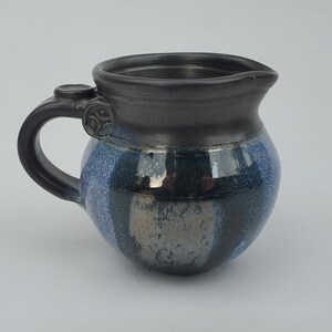 May include: A small, dark blue and black ceramic creamer with a handle. The creamer has a metallic glaze and a striped pattern.