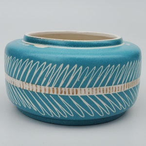 May include: A turquoise ceramic bowl with a cream-colored interior. The exterior features a white, diagonal, feather-like pattern above a gold band. The bowl has a rounded shape and a slightly textured surface.