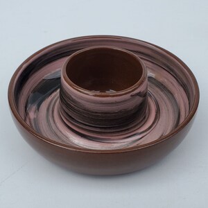 May include: A brown and pink ceramic egg cup with a matching saucer. The cup and saucer have a swirl pattern in black, pink, and brown.