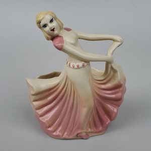 May include: A ceramic figurine of a woman in a pink and white dress, holding her skirt with one hand and her other arm outstretched. The figurine is a planter with a hollow space in the skirt.