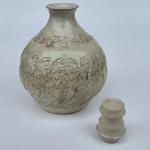 May include: A light beige ceramic decanter with a rounded body and a narrow neck. The surface features a textured, carved pattern around the middle section. A separate, matching tiered ceramic stopper sits beside the decanter on the surface.