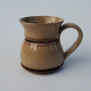 May include: A brown ceramic mug with a wide base and a flared rim. The mug has a brown glaze with a lighter brown band around the middle.