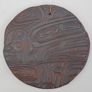 May include: A round, brown ceramic wall hanging with a carved design of stylized animal figures. The design is reminiscent of Northwest Coast Indigenous art.