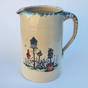 May include: A cream-colored ceramic pitcher with a decorative garden scene featuring birdhouses, a white picket fence, and flowers. The rim and handle have a green and blue speckled design.