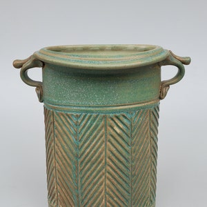 May include: A green ceramic vase with two handles and a herringbone pattern. The vase is glazed in a light green color with a brown glaze on the herringbone pattern.