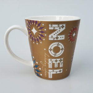 May include: A gold ceramic mug with a white handle. The mug has a festive design with the word "NOEL" in a stylized font and a pattern of fireworks. The text "holiday cheer" is written in cursive on the side of the mug.