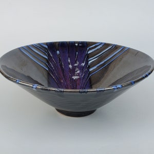 May include: A large, dark gray ceramic bowl with a wide rim and a flared base. The bowl has a purple and blue glaze with streaks of white.