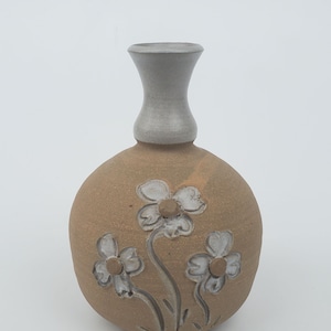 May include: A small, brown ceramic vase with a white glaze on the neck. The vase is decorated with three white flowers with brown centers.