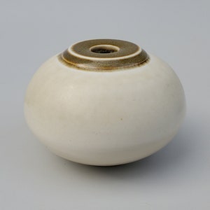 May include: A white ceramic vase with a brown rim. The vase is round and has a small opening at the top.