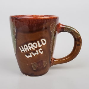 May include: A brown ceramic mug with a white glaze and the words "Harold WWC" written on the side.