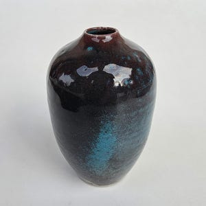 May include: A ceramic vase with a glossy finish, featuring a deep dark purple and black base color that transitions into a vibrant, mottled turquoise blue patch on the front. The vase has a rounded body and a narrow, circular opening at the top.