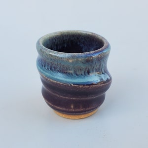 May include: A small, hand-thrown ceramic cup with a blue and purple glaze. The cup has a slightly uneven, organic shape.