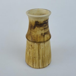 May include: A ceramic vase with a cylindrical shape and a wide opening. The vase features a textured, vertical design on the lower portion and a darker brown band around the middle. The color palette includes shades of tan and brown.
