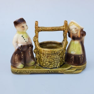 May include: A ceramic wishing well planter with two figurines, a boy and a girl, standing on either side. The well is green and brown with a stone-like texture. The base of the planter reads "Wishing Well".