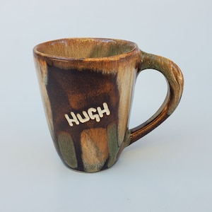 May include: A brown and green ceramic mug with a white glaze. The mug has a handle and the word "HUGH" written on the side.