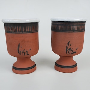May include: Two terracotta cups with white rims and black painted designs. The cups have a wide base and a narrow top. The cups are signed "Hisa" in black ink.