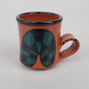 May include: A small, terracotta mug with a black rim and a green and black geometric design. The mug has a handle.