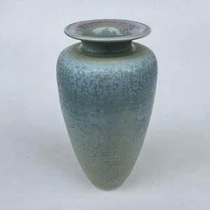 May include: A tall, ceramic vase with a wide, flat rim and a textured surface. The vase has a gradient of colors, transitioning from a light green at the base to a speckled blue-gray at the top. The rim is a darker, speckled brown.