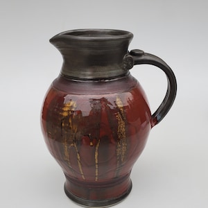 May include: A red and brown ceramic pitcher with a dark brown glaze and a handle. The pitcher has a rounded body and a wide mouth.