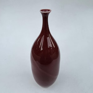May include: A tall, burgundy-colored vase with a slender neck and a rounded body. The vase has a glossy finish and a subtle, swirling pattern. The rim of the vase is a lighter shade of burgundy. This decorative vase is suitable for home decor.