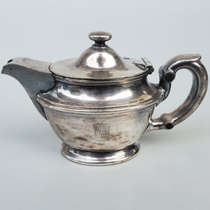 May include: Silver teapot with a lid and a handle. The teapot has an ornate design and a crest on the front.
