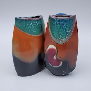 May include: Two ceramic vases with a unique, abstract design. The vases are orange, black, green, and red with a glossy finish.