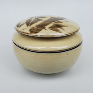 May include: A ceramic jar with a lid, featuring a brown and white leaf design. The jar has a cream-colored body with a blue stripe around the rim.