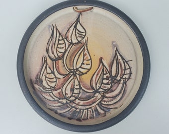 May include: A ceramic wall hanging with a black rim and a beige background. The design features a stylized floral pattern in shades of brown, beige, and white.