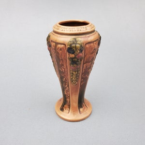May include: A vintage, peach-colored vase with a flared base and a detailed, textured design. The vase features embossed grape clusters and foliage, with a dark brown and green color palette. The top rim has a decorative band.