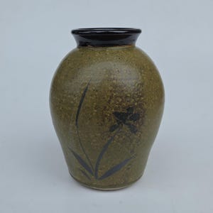 May include: A handcrafted ceramic vase with a speckled olive green glaze. The vase features a dark brown rim and a black floral design. The vase has a rounded body and a slightly flared opening, suitable for displaying flowers or as a decorative piece.