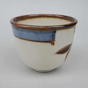 May include: A white ceramic teacup with a brown rim and a blue and brown stripe design. The cup has a simple, elegant design.