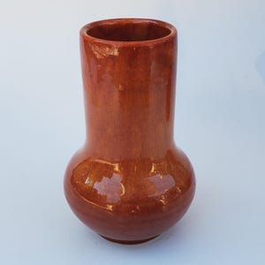 May include: A glossy, reddish-brown ceramic vase with a bulbous base and a cylindrical neck. The vase has a smooth, reflective surface, and the opening is circular. The design is simple and elegant, suitable for displaying flowers or as a decorative piece.