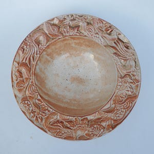 May include: A handmade ceramic plate with a light brown center and a detailed, raised floral and dragon design around the rim. The plate has a rustic, earthy aesthetic, perfect for serving food or as a decorative piece.
