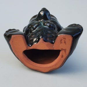 May include: A black ceramic figurine of a bear lying on its back with its mouth open. The bear is glazed in a dark green color and has a brown clay interior.
