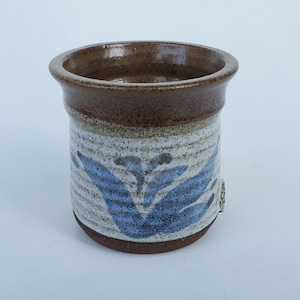 May include: A handmade ceramic vase with a cylindrical shape. The top rim is dark brown, transitioning to a speckled white and light brown body. The vase features a blue floral design. The base is a darker brown color.