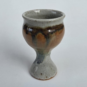 May include: A ceramic goblet featuring a speckled light gray base and rim, with a central band of warm brown glaze that drips downward into dark, moody streaks. The vessel has a rounded bowl, a narrow stem, and a wide, stable circular foot.