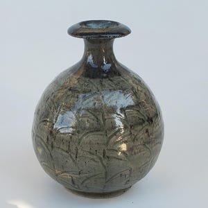 May include: A handcrafted ceramic vase with a rounded body and a narrow neck. The vase is a muted gray-green color with a textured, patterned surface. The rim of the vase is dark gray. The vase is suitable for home decor.