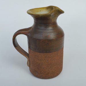 May include: A handmade ceramic pitcher with a handle. The pitcher has a brown and tan color scheme. The top of the pitcher has a spout for pouring. The pitcher is a rustic, handcrafted piece.