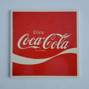 May include: Square red and white Coca-Cola sign. The sign features the Coca-Cola logo in white script with the word "Enjoy" above it. The words "Trade-mark ®" are below the logo.