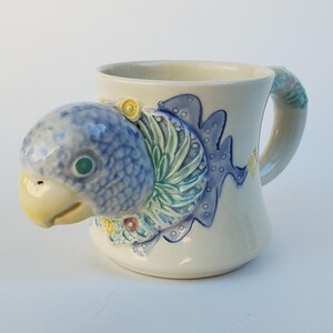 May include: A white ceramic mug with a blue and green parrot design. The parrot's head is sculpted and extends from the mug's handle.