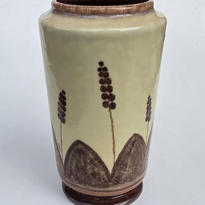 May include: A cylindrical ceramic vase with a glossy cream-colored glaze. The base and rim are finished in a dark brown glaze. The body features hand-painted dark brown illustrations of three stylized floral stalks rising from rounded hills.