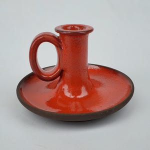 May include: A red ceramic candle holder with a handle. The holder has a textured surface and a brown base.