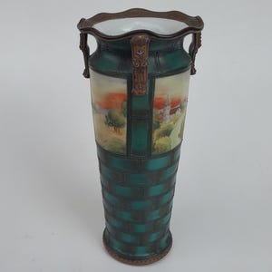 May include: An antique, tall vase with a green brick-like pattern on the lower portion and a landscape painting on the upper portion. The vase has a brown rim and handles, and a white interior.